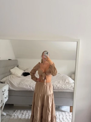 Beige set med lång kjol och topp - Säljer ett beige set med lång kjol och matchande topp med puffärmar och knytning framtill. Setet är tillverkat i ett mjukt, vävt material och har en avslappnad, bohemisk stil. Perfekt för dig som gillar trendiga och bekväma outfits.