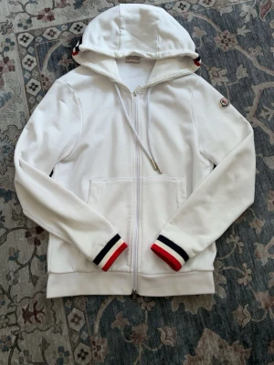 Moncler hoodie - Säljer denna Moncler hoodien i vit färg då den inte passar mig längre. Perfekt nu inför sommaren för dig som vill vara stilig. Den har inga skador eller slitage. Hör av er vid frågor !