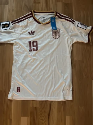 Vit Spanien fotbollströja Adidas - Säljer en vit Spanien fotbollströja från Adidas med vinröda detaljer, nummer 19 och namnet Lamine Yamal på ryggen. Tröjan har officiella klubbmärken, broderad Adidas-logga och bruna ränder på axlarna. Tillverkad i ett lätt och svalt material, perfekt för match eller träning.