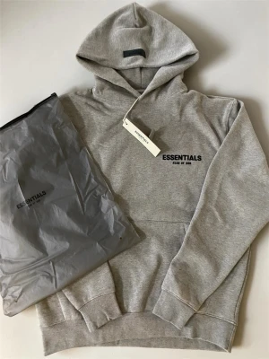 Grå Essentials Fear of God hoodie - Snygg grå hoodie från Essentials Fear of God med svart logga på bröstet. Klassisk passform med huva och känguruficka framtill. Tillverkad i mjukt bomullsmaterial som känns skönt mot huden. Perfekt för en avslappnad streetwear-look.