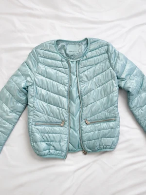 Ljusblå pufferjacka från Amisu - Snygg ljusblå pufferjacka från Amisu med rund halsringning och dragkedja framtill. Jackan har två framfickor med dragkedja och quiltad design i ett glansigt material. Perfekt för kalla dagar och ger en trendig look.