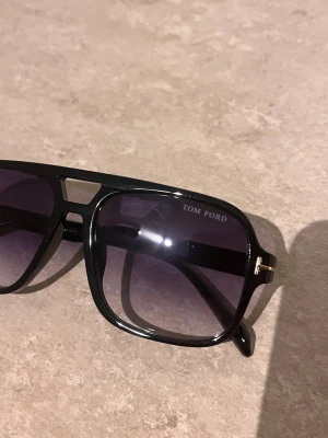 Svarta solglasögon från Tom Ford - Snygga svarta solglasögon från Tom Ford med klassisk pilotform och mörka glas. Bågarna har gulddetaljer vid gångjärnen och en cool design med en öppning ovanför näsryggen. Perfekta för att lyfta din stil till nästa nivå.