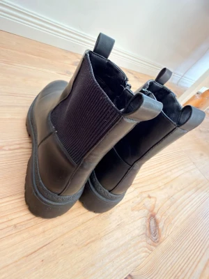 Svarta chunky Chelsea boots - Säljer ett par svarta Chelsea boots med chunky sula och elastiska paneler på sidorna. Skorna har dragflikar både fram och bak för enkel påtagning. Tillverkade i slätt konstläder som ger en clean och modern look.