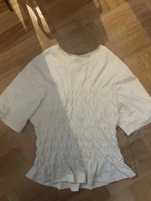 Vit smockad topp från Gina Tricot - Supersöt vit topp från Gina Tricot i storlek XS. Toppen har smockad midja, korta ärmar och en liten volangkant nedtill. Perfekt att styla med jeans eller kjol för en fräsch look.