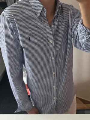 Blårandig skjorta från Ralph Lauren - Klassisk blårandig skjorta från Ralph Lauren med button-down krage och broderad logga på bröstet. Skjortan har långa ärmar, vita knappar och är tillverkad i bomull. Perfekt för en clean och stilren look.