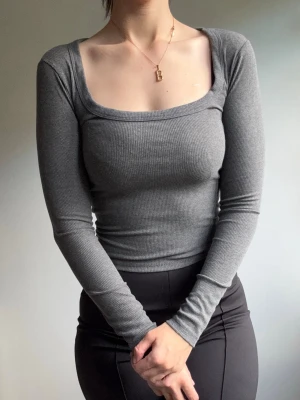 Grå ribbad långärmad topp - Säljer en stilren grå ribbad topp med lång ärm och fyrkantig urringning. Toppen är tight i modellen och har en mjuk, stretchig känsla som sitter snyggt på kroppen. Perfekt att matcha med både jeans och kjol.