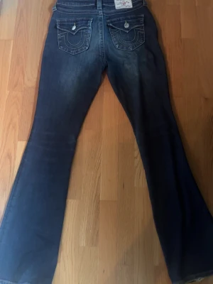 Blå bootcut jeans från True Religion - Säljer ett par mörkblå bootcut jeans från True Religion med ikoniska fickor bak och kontrastsömmar. Jeansen har medel midja och klassisk femficksdesign. Perfekta för dig som gillar en avslappnad men ändå snygg stil.