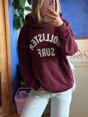 Vinröd hoodie från Hollister - Vinröd hoodie från Hollister med vit text. Den har en dold ficka i sidan med dragkedja vilket är smidigt. 