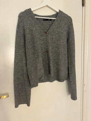 Grå stickad kofta med knappar - Mysig grå stickad kofta med v-ringning och två bruna knappar framtill. Koftan har lång ärm och avslappnad passform, perfekt att slänga över en t-shirt. Ribbstickade muddar vid ärmslut och nederkant ger en enkel men stilren look.