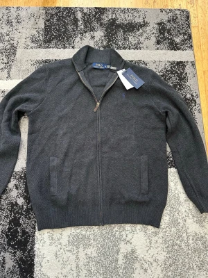Grå ziptröja från Polo Ralph Lauren - Stilren grå ziptröja från Polo Ralph Lauren med hög krage och diskret broderad logga på bröstet. Tröjan har två fickor framtill, ribbade muddar och är tillverkad i mjukt stickat material. Perfekt för en avslappnad men snygg look. OBS LITEN I STORLEKEN PASSA STORLEK L