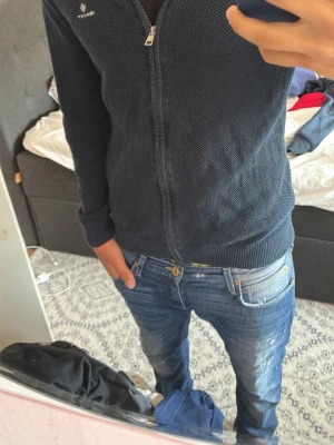 Mörkblå ziptröja från Gant - Mörkblå ziptröja från Gant med diskret struktur i materialet. Tröjan har hel dragkedja framtill, ribbade muddar vid ärmslut och nederkant samt en liten logga på bröstet. Perfekt för en avslappnad och stilren look.