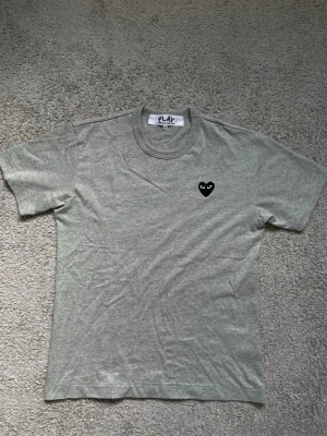 Grå t-shirt Comme des Garçons Play - Snygg grå t-shirt från Comme des Garçons Play med det ikoniska svarta hjärtat broderat på bröstet. Klassisk passform och rund halsringning. Perfekt basplagg för en clean och stilren look.