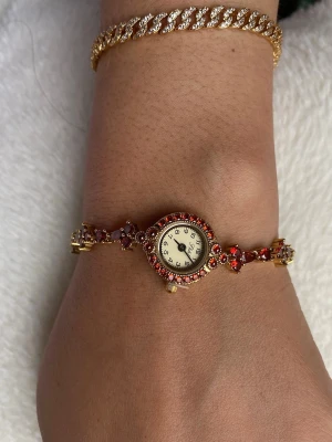 Guldig guldpläterad  armbandsklocka med zirikon stenar - Unik armbandsklocka i guldton med röda vinröda stenar. Klockan har ett runt ur med dekorativa detaljer runt boetten och ett smyckat armband med blommönster. Perfekt för dig som vill ha något extra till din stil.