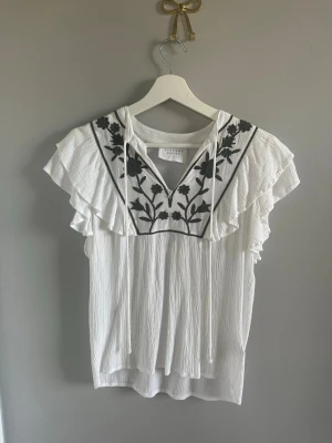 Vit blus med volang och broderi - Säljer en luftig vit blus från Part Two med svarta broderade blommor på bröstet och volangdetaljer längs axlarna. Blusen har v-ringning med knytband och är ärmlös, perfekt för en trendig och avslappnad look.
