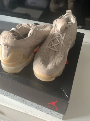 Beige Nike Air Vapormax sneakers - Säljer ett par beige Nike Air Vapormax sneakers med stickad ovandel och genomskinlig sula. Skorna har en vit swoosh på sidan och elastiska snören för en clean look. Perfekta för dig som gillar streetwear och vill ha något bekvämt och stilrent.