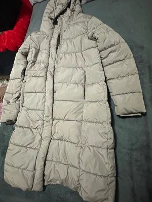 Lång beige dunjacka med huva - Lång och varm beige dunjacka med huva och quiltad design. Jackan har raka ärmar, dold dragkedja och tryckknappar framtill. Perfekt för kalla vinterdagar och ger en stilren look.