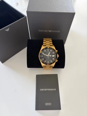 Emporio Armani guld herrklocka 100% äkta  - Stilren klocka från Emporio Armani med guldfärgad länk i metall och svart urtavla. Klockan har kronograf, datumvisning och tydliga indexmarkeringar. Kommer i originalbox med garanti. Perfekt accessoar för dig som vill sticka ut.