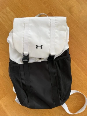 Svartvit ryggsäck från Under Armour - Snygg svart och vit ryggsäck från Under Armou. Ryggsäcken har två stora meshfickor på sidorna, justerbara axelband och spännen framtill. Tillverkad i slitstarkt syntetmaterial och har en modern, sportig design. Väl omhändertagen och i bra skick.