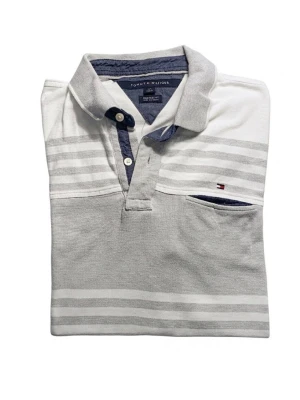 Randig pikétröja från Tommy Hilfiger - Snygg pikétröja från Tommy Hilfiger i vitt och ljusgrått med horisontella ränder. Tröjan har klassisk krage, knappar vid halsen och en liten bröstficka med logga. Perfekt för en avslappnad och stilren look.