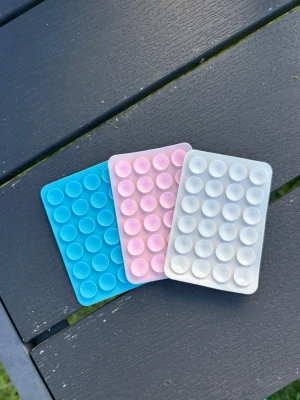 Mobilgrepp Pop it fidget-leksaker i silikon - Tre mobilgrepp med klister på baksidan. Nya.  rektangulära pop it fidget-leksaker i silikon, en blå, en rosa och en vit. Varje leksak har ett rutnät med bubblor att trycka på för stresslindring eller tidsfördriv. Perfekt att ha i väskan eller på skrivbordet.