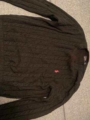 Svart kabelstickad tröja Polo Ralph Lauren - Svart kabelstickad tröja från Polo Ralph Lauren med rund halsringning och röd broderad logga på bröstet. Tröjan har ribbade muddar vid ärmslut och nederkant. Perfekt för en stilren och klassisk look.