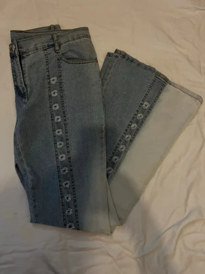 Blå jeans med blommönster längst benen  - Säljer ett par ljusblå jeans med utsvängda ben och blommönster längs frambenen. Jeansen har klassisk femficksdesign och medium midja. Perfekta för dig som gillar en unik och trendig look.