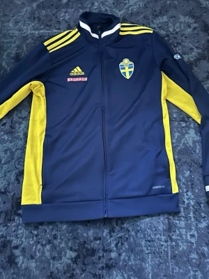 Blå-gul Adidas Sverige träningsjacka - Säljer en blå träningsjacka från Adidas med gula detaljer och Sveriges landslagsemblem på bröstet. Jackan har hel dragkedja, gula ränder på axlarna och Bauhaus-logga. Tillverkad i funktionsmaterial, perfekt för träning eller sport.