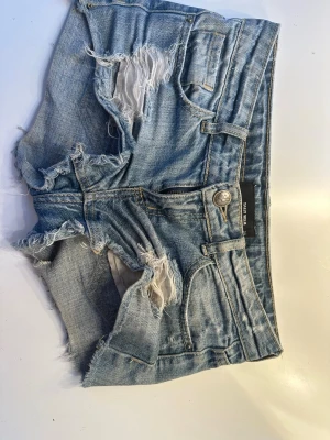 Blå slitna jeansshorts  - Säljer ett par blå jeansshorts med slitna detaljer och råa kanter. Shortsen har klassisk femficksdesign, knappgylf och är tillverkade i mjuk bomull. Perfekta för varma dagar och ger en avslappnad look. 