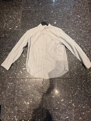 Randig skjorta från Ralph Lauren - Klassisk vit skjorta med tunna blå ränder från Ralph Lauren. Skjortan har lång ärm, knappar framtill och en liten broderad logga på bröstet. Perfekt för en stilren look och passar till många olika outfits. Inte ink frakt 