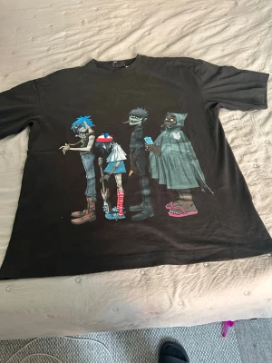 Svart Gorillaz t-shirt med tryck - Cool svart t-shirt med stort färgglatt Gorillaz-tryck på framsidan. Klassisk passform och rund hals. Perfekt för dig som gillar bandmerch och streetwear. Materialet känns mjukt och skönt mot huden.