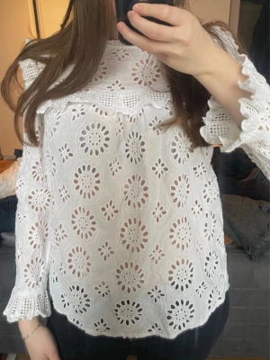 Vit broderad blus med volangdetaljer - Säljer en vit blus med broderade blommönster och volangdetaljer vid axlar och ärmslut. Blusen har lång ärm och är tillverkad i ett luftigt, genombrutet tyg som ger en skön känsla. Perfekt för dig som gillar en feminin och trendig stil.                                                                           Defekt: är tyvärr ett hål under ena ärmen/armhålan men syns inte väl & går nog bra att laga💕