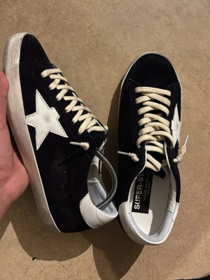 Golden Goose  - Snygga svarta sneakers med vit stjärndetalj på sidan och vita partier vid häl och sula. Skorna har vita snören och en klassisk låg siluett. Ovandelen verkar vara i mocka och detaljerna i läder. Perfekta för en trendig streetstyle-look. Skorna är designade för att de mörkblåa och svarta ut, när du har på dig svarta kläder dem kommer de svarta ut. När du har på dig mörkblåa eller ljus kläder kommer de se mörkblåa ut.