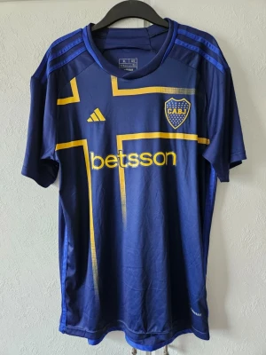 Blå Boca Juniors matchtröja Adidas - Snygg blå Boca Juniors matchtröja från Adidas med gula detaljer och klubbmärke på bröstet. Tröjan har korta ärmar, rund hals och tryckt sponsorlogga framtill. Perfekt för fotbollsträning eller att bära som supporter. (Obs r3plika)