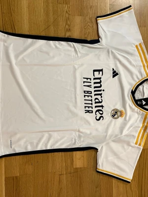 Real Madrid matchtröja  - Säljer en vit Real Madrid matchtröja från Adidas med svarta och gula detaljer. Tröjan har klubbmärke och Adidas-logga på bröstet samt 'Emirates Fly Better' tryckt framtill. Tillverkad i ett lätt och ventilerande material, perfekt för fotbollsträning.    Fraktar samma eler dagen efter du köpt!!! Endast en kvar i lager!!!