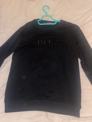 Svart sweatshirt från Hugo Boss - Säljer en svart sweatshirt från Hugo Boss med stor broderad logga på bröstet. Tröjan har rund halsringning och långa ärmar. Materialet känns mjukt och bekvämt, perfekt för en avslappnad stil. Passar dig som gillar klassiska och stilrena plagg.