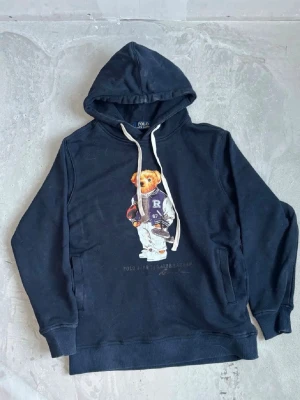 Svart hoodie med Polo Bear tryck - Svart hoodie med huva och vita dragsnören från Polo Ralph Lauren. På bröstet finns ett stort tryck av den ikoniska Polo Bear i sportig outfit. Hoodien har känguruficka och ribbade muddar. Perfekt för en avslappnad och cool stil.