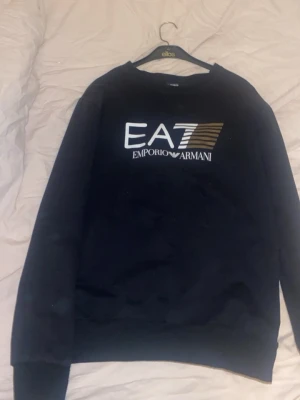 Svart sweatshirt EA7 Emporio Armani - Svart sweatshirt från EA7 Emporio Armani med tryckt logga i vitt och guld på bröstet. Klassisk rund halsringning och långa ärmar. Tillverkad i mjukt material för en avslappnad och stilren look.