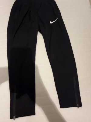 Svarta träningsbyxor från Nike - Säljer ett par svarta träningsbyxor från Nike med vit logga på låret. Byxorna har dragkedjor vid bensluten och elastisk midja för skön passform. Perfekta för löpning eller gym. Materialet känns lätt och smidigt. Mycket bra för sommaren. 