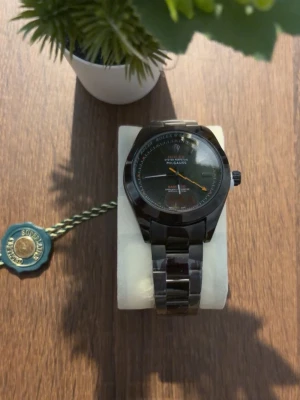 Klocka  - Stilren svart klocka med metallarmband och rund urtavla. Urtavlan har detaljer i orange och grönt samt tydliga markeringar. Perfekt accessoar för dig som vill ha en klassisk men modern look.