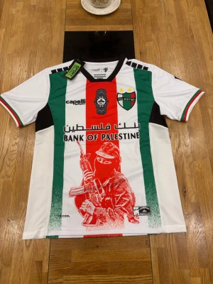 Palestino matchtröja med tryck - Säljer en officiell Palestino matchtröja från Capelli Sport. Tröjan har gröna, röda och vita vertikala ränder, klubbmärke på bröstet och flera sponsorloggor. Unikt tryck framtill med motiv och texten 'Bank of Palestine'. Korta ärmar och rund hals.