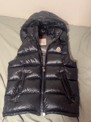 Svart dunväst med huva från Moncler - Snygg svart dunväst från Moncler med glansig finish, huva och två dragkedjeförsedda fickor. Västen har Moncler-logga på bröstet och quiltad design som ger en modern look. Perfekt för lager-på-lager-stil under kyliga dagar.