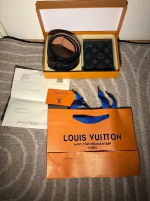 Svart Louis Vuitton bälte i skinn - Säljer ett svart bälte från Louis Vuitton i skinn med klassiskt LV-spänne. Bältet har en stilren design och kommer med en matchande plånbok i svart med monogrammönster. Perfekt accessoar för dig som gillar lyxiga detaljer.