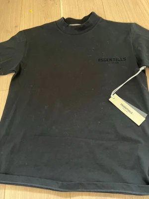 Svart Essentials t-shirt med hög krage - Säljer en svart t-shirt från Essentials med hög krage och diskret logga på bröstet. T-shirten har en clean och minimalistisk look, perfekt för dig som gillar streetwear. Tillverkad i mjukt bomullsmaterial och har en relaxed fit.