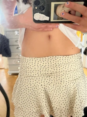 Så fin Vit kortkjol med svarta prickar - Supersöt vit kortkjol med svarta prickar och volangkant. Kjolen har en hög midja och är gjord i ett lätt och mjukt material som känns skönt mot huden. Perfekt för varma dagar och ger en riktigt fräsch look.