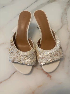 virala mules från H&M  - Säljer ett par vita pumps med öppen tå och låg klack. Skorna har en snygg ovandel täckt av skimrande paljetter i vitt och pärlemor. Klacken är smal och ger en elegant look. Perfekta för dig som vill sticka ut lite extra. Helt slutsålda och jätte svåra få tag på 