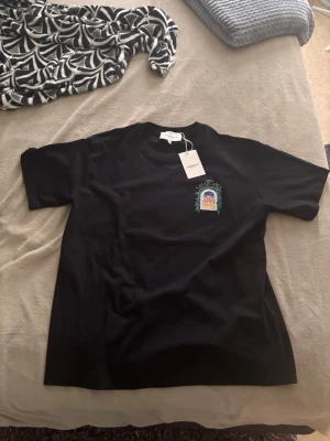 Svart t-shirt med tryck Casablanca - Snygg svart t-shirt med färgglatt tryck på bröstet och stort motiv på ryggen med texten 'Avenida Casablanca' och ett fönster med gröna blad och byggnad. Klassisk passform och rund hals. Perfekt för dig som gillar streetwear och grafiska prints.