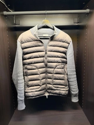 Moncler cardigan - Snygg grå dunväst från Moncler med stickade ärmar och quiltad front. Västen har dragkedja framtill, två fickor med dragkedja och en klassisk Moncler-logga på ärmen. Perfekt för lager-på-lager och streetstyle.