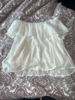Vit offshoulder blus med volang - Säljer en söt vit offshoulder blus med volangdetalj upptill. Blusen är ärmlös och har ett tunt, lätt och luftigt material som passar perfekt till varmare dagar. Enkel att styla med både jeans och kjol.