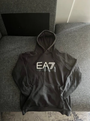 Svart hoodie EA7 Emporio Armani - Snygg svart hoodie från EA7 Emporio Armani med stor logga på bröstet. Klassisk modell med huva och dragsnören samt en stor magficka. Perfekt för en avslappnad och trendig stil. Mjuk bomullskänsla och ribbade muddar vid ärmar och nederkant.