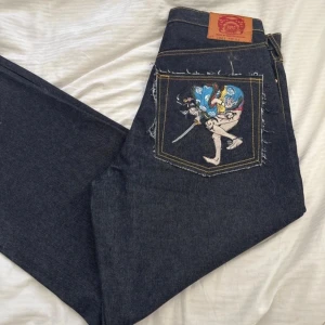 Jeans med print - Ej använda unika mörkblå jeans med raka ben och coola broderade prints på bakfickorna. Tjock denim med kontrastsömmar och fransade fickkanter.
